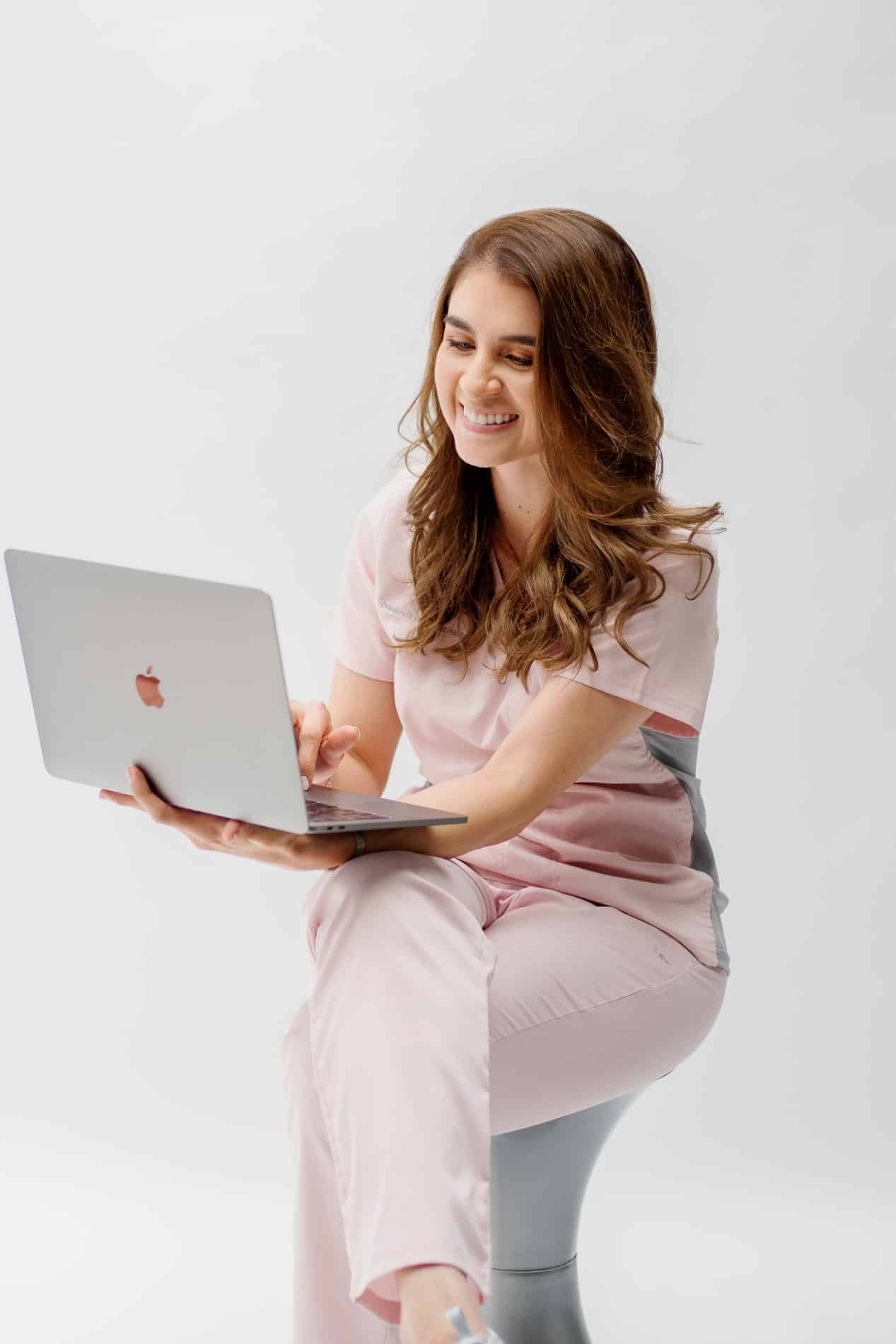 Dra. Natalia Morales sonriente y sentada en un banco usando su Macbook Pro bajo un fondo blanco