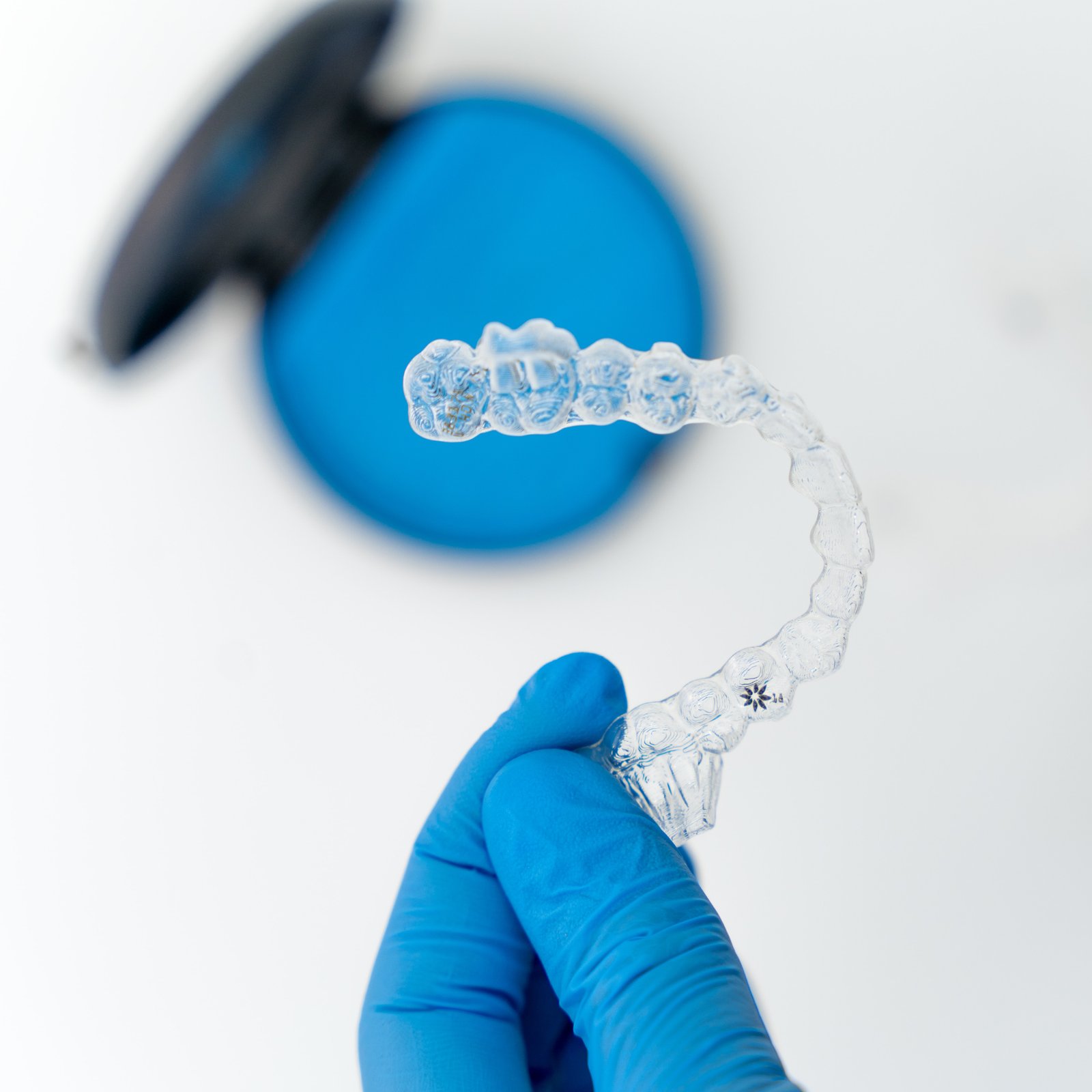 Mano con guante azul sosteniendo un alineador de Invisalign con un estuche en el fondo